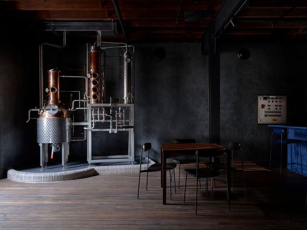 Dark industrial-style interior featuring a prominent copper and stainless-steel distillation apparatus along the left wall, exposed timber ceiling structure, black-painted structural beams, charcoal walls, and dark timber flooring. A small dining or tasting table with black chairs occupies the centre, while a blue counter or bar area and control panel are visible at right. The space resembles a boutique distillery tasting room or production showroom.