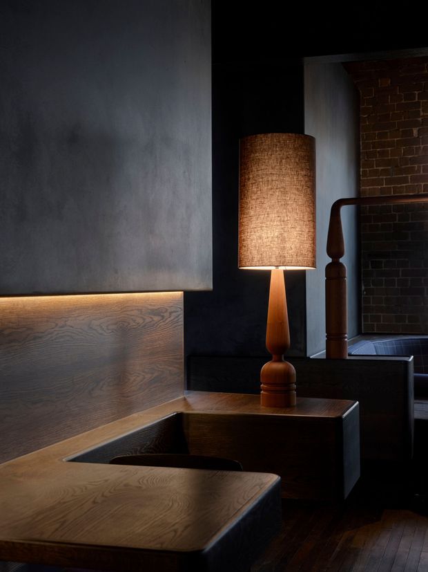 A dark, moody interior vignette featuring sculptural dark timber built-in seating or cabinetry with a broad wood-grain surface, a warm glowing table lamp with a woven cylindrical shade, charcoal plaster walls, and exposed dark brickwork in the background. A dark timber stair or balustrade element is visible to the right, contributing to the industrial, refined atmosphere.