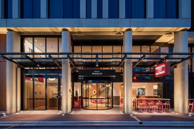 Night-time exterior view of a contemporary hotel entrance beneath a large commercial canopy. The facade features blue vertical cladding panels, extensive floor-to-ceiling glazing, cylindrical columns, a glass-and-metal porte-cochère, and illuminated signage for Hotel Indigo and a restaurant or bar. Outdoor seating is visible along the frontage.