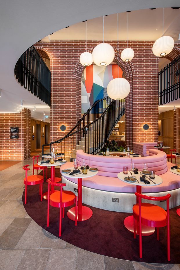 A vibrant hospitality interior with a circular pink upholstered banquette surrounding a central dining area, paired with bright red chairs and small round tables. The space features exposed red brick walls, a sweeping black metal staircase and balustrade, colourful abstract wall artwork, and multiple ribbed globe pendant lights. Grey stone-look floor tiles and dark burgundy carpet define the seating area, creating a playful, theatrical restaurant or bar atmosphere.