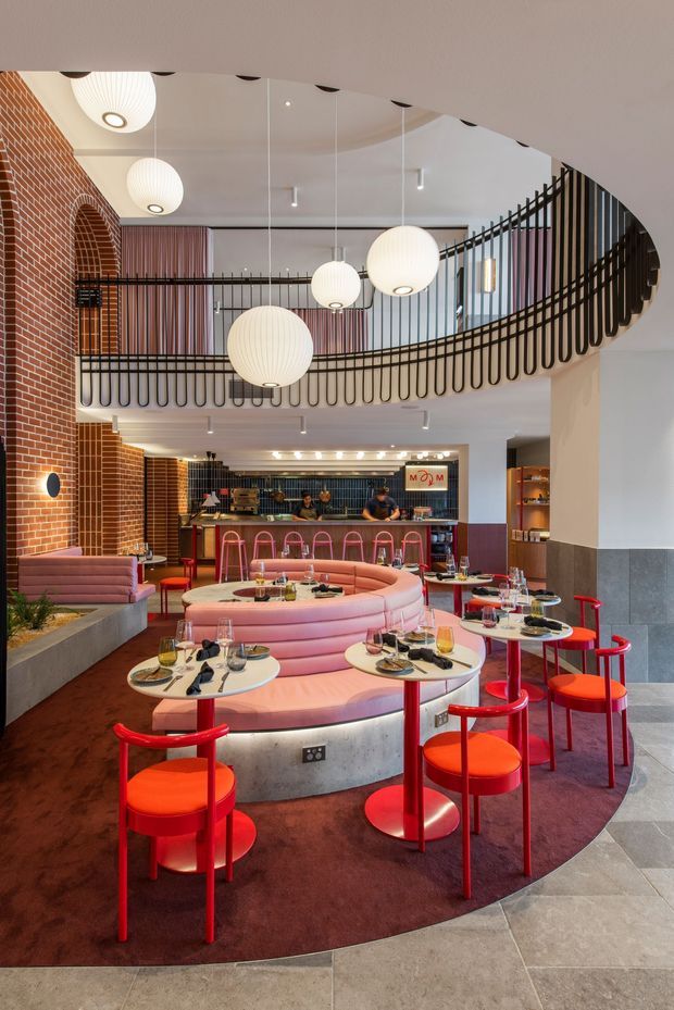 A vibrant multi-level restaurant interior with a curved mezzanine and black metal balustrade overlooking a central dining area. The space features exposed red brick arches, a white sculptural ceiling, ribbed globe pendant lights, pink upholstered curved banquettes, red dining chairs and tables, a bar at the rear, and dark burgundy carpet with light stone-look floor tiles. The overall design is playful, bold, and contemporary with strong pink and red accents.