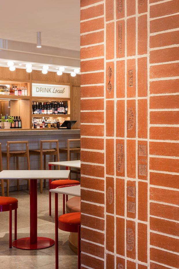 Interior view of a contemporary bar or casual dining venue. A prominent red-orange exposed brick partition with pale mortar dominates the foreground, while a timber drinks bar and shelving display bottles beneath a “DRINK Local” sign. White rectangular tables, red pedestal bases, and rounded red and tan stools occupy the seating area. Warm integrated lighting and a neutral tiled floor create a lively hospitality atmosphere.