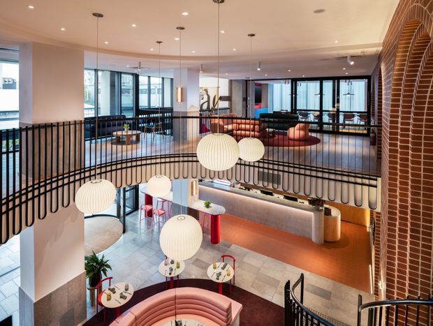 A multi-level contemporary commercial interior, likely a hospitality or coworking space, viewed from an upper landing. The open atrium features curved black metal balustrades, multiple spherical pendant lights, lounge seating, small round tables, a long illuminated counter, expansive glazing, and a prominent curved red-brick feature wall with arched detailing. The palette combines warm terracotta and blush upholstery with pale stone flooring, white walls, black metalwork, and timber-toned accents.