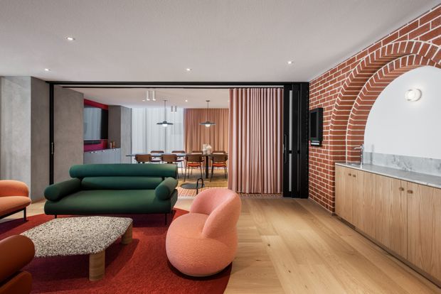 Warm, eclectic open-plan living and dining interior with a dark green rounded sofa, sculptural peach lounge chairs, textured coffee table, and red rug. A timber floor runs through the space, while a striking exposed red-brick arched feature wall frames a marble-topped timber vanity or bar-like joinery. Black-framed glazed sliding doors, vertical timber screening, pendant lights, and a partially visible kitchen complete the layered contemporary fit-out.