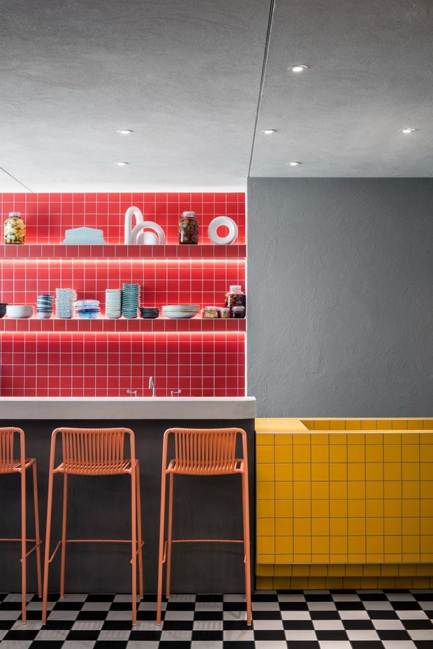 Bold, highly graphic kitchen or bar interior with a black-and-white checkerboard floor, vivid red square-tile backsplash and open shelving, a dark island with a pale benchtop, and a mustard-yellow tiled wall. Coral-orange woven bar stools sit at the island. The ceiling and adjacent wall are textured grey with recessed downlights, creating a playful retro-contemporary atmosphere.