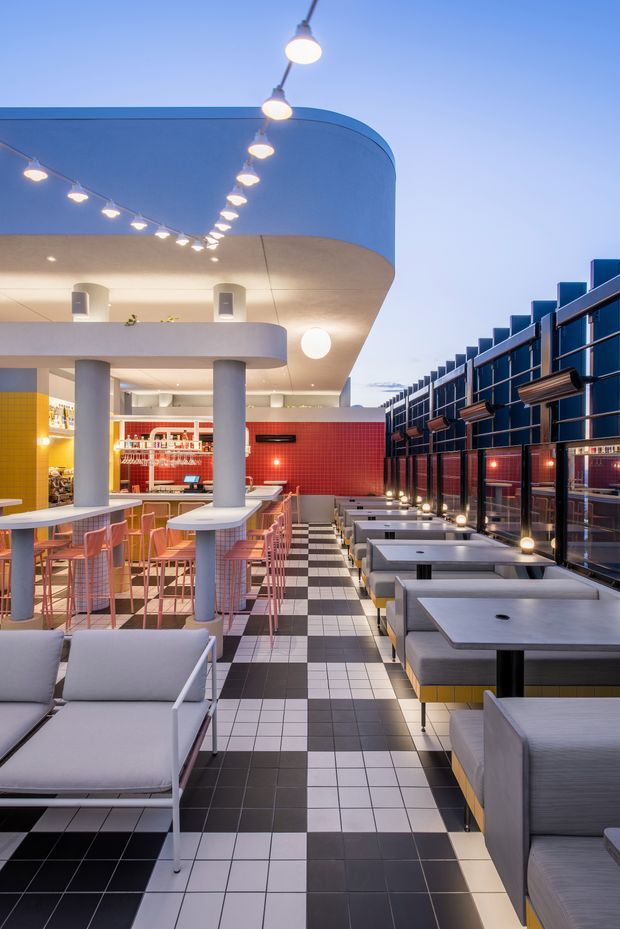 A stylish commercial dining and lounge interior, likely a restaurant or hospitality venue, photographed at dusk. The space features a dramatic curved white ceiling and mezzanine-like balcony, exposed round columns, suspended globe lighting, a red tiled service/bar wall, yellow tiled accents, and extensive outdoor or glazed-edge booth seating. Black-and-white checkerboard floor tiles create a bold graphic pattern, while pink bar stools and upholstered grey seating add colour and comfort. A dark metal glazed screen lines the right side.