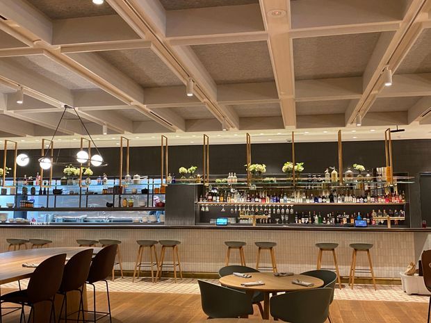 A sophisticated contemporary commercial bar and dining space with a long tiled bar counter, extensive back-bar shelving stocked with bottles and glassware, pendant globe lights, and flexible café-style seating. The ceiling features a prominent coffered timber-look grid with acoustic infill panels, while dark wall finishes contrast with warm timber flooring and neutral furnishings.