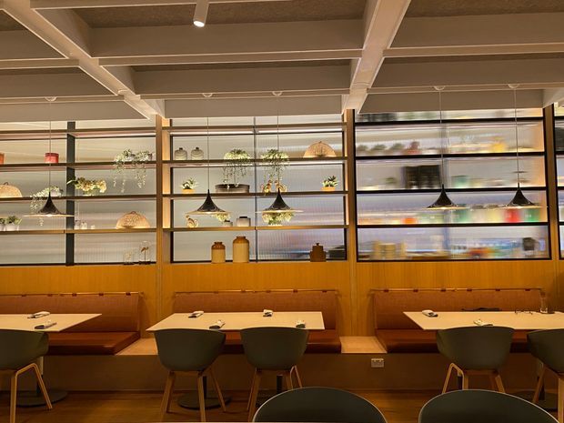 A warm, contemporary restaurant or café dining interior with timber booth seating, small dining tables, upholstered chairs, pendant lights, and open shelving displaying plants, ceramics, and glassware. The space features exposed structural ceiling beams, timber wall panelling, dark metal shelving frames, and a muted earthy palette.