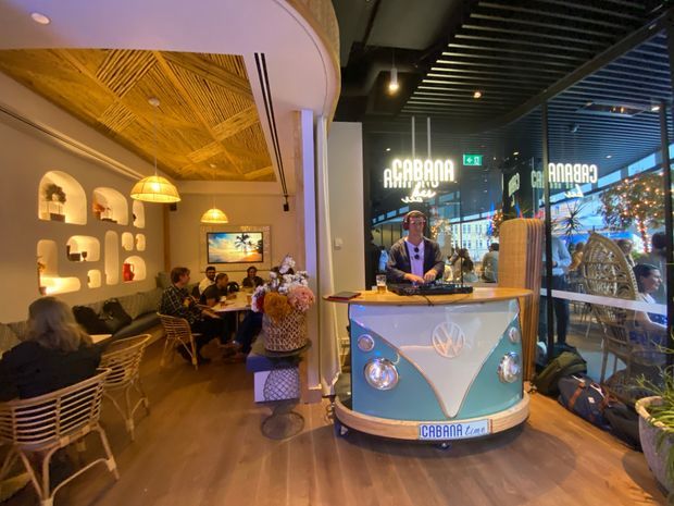 A warmly lit retro-themed commercial hospitality interior, likely a café or bar. The foreground features a turquoise vintage Volkswagen van-style counter, while the seating area includes wicker chairs, small tables, a dark upholstered banquette, decorative illuminated wall niches, pendant lights, and a distinctive woven timber ceiling. Large glazed frontage opens visually to a brightly lit shopping or urban environment outside.