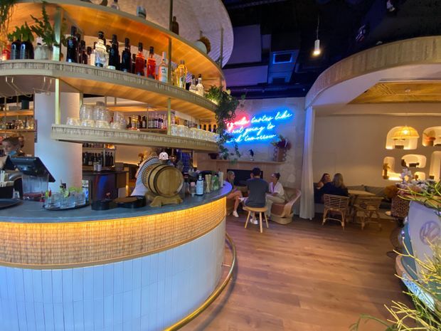 A warmly lit hospitality interior featuring a curved bar with illuminated tiled frontage, layered open shelving stocked with bottles and glassware, integrated timber barrel detailing, and an intimate lounge seating area. The space has a dramatic dark ceiling, arched architectural forms, timber flooring, pendant lighting, indoor planting, and a bright neon wall sign, creating a vibrant, atmospheric cocktail-bar setting.