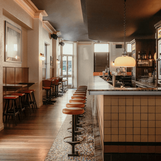 A warmly lit, narrow bar interior with a long tiled counter, a row of rust-coloured swivel stools, espresso equipment, glassware shelving, dark timber flooring, framed wall artwork, and tall windows. The space has a refined vintage-inspired hospitality design with contrasting dark ceiling and warm neutral finishes.