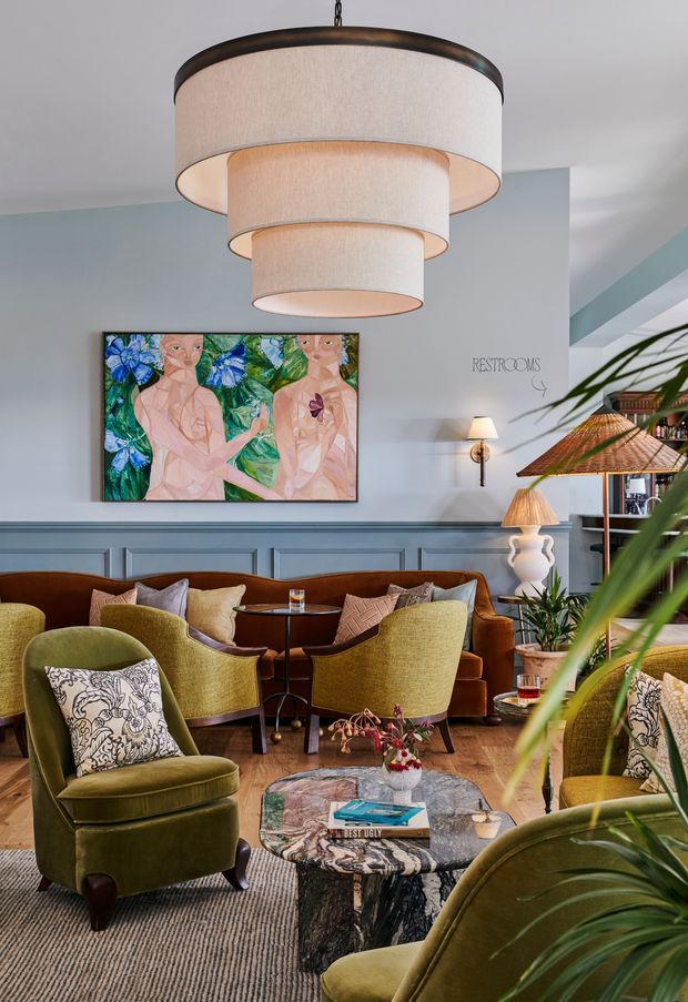 A richly layered lounge or hotel-style sitting room with olive green upholstered armchairs, rust and brown seating, patterned cushions, a sculptural stone coffee table, timber flooring, and a large tiered fabric pendant light. Pale blue walls feature decorative wall panelling, a colourful floral artwork, and signage reading “RESTROOMS”; tropical plants and a timber-framed glazed opening appear at the right. The room has an eclectic, refined, warm character with strong botanical and vintage-inspired detailing.