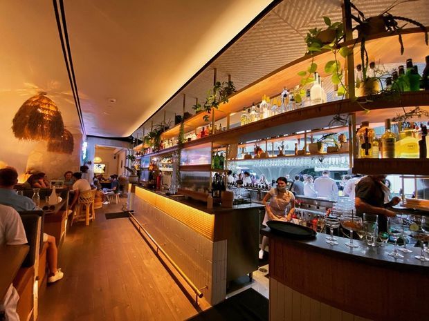 A warmly lit contemporary restaurant and cocktail bar interior, viewed along a long central bar. The space features extensive illuminated bottle and glassware shelving, timber-toned bar cabinetry, a dark wood floor, hanging plants, pendant lighting, and dining seating along the left side. Staff and patrons are visible behind and around the bar.