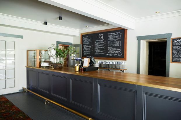 A refined hospitality counter or bar interior featuring dark navy/charcoal panelled fixed cabinetry, a warm timber countertop, chalkboard menu boards, brass foot rail, and pale walls with decorative ceiling mouldings. The space has a traditional yet contemporary pub or café character, with a service area and doorway in the background.
