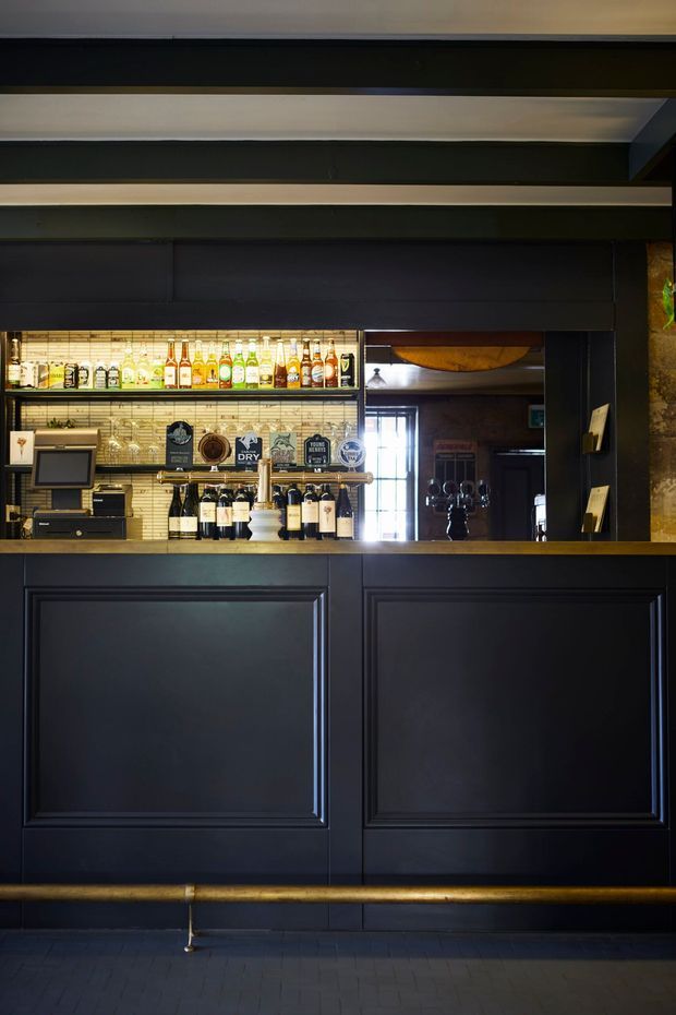 A dark, sophisticated bar or reception counter with deep navy-black raised-panel cabinetry, brass-toned rail and trim, a mirrored backbar, illuminated shelves of bottles and glassware, and black-painted ceiling beams. The space has a moody, refined hospitality interior with warm metallic accents and tiled flooring.