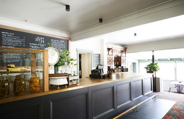 Bright, refined café or hospitality counter with a long dark paneled service bar, warm timber countertop, chalkboard menu wall, glass pastry display, coffee equipment, and shelving. The space has white coffered detailing, recessed and cylindrical lighting, and large glazed openings that create an airy atmosphere.