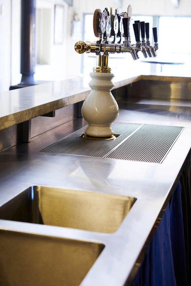 Close-up of a commercial bar service counter featuring a multi-tap beer dispenser mounted on a decorative ceramic pedestal. The bar has a reflective stainless-steel worktop, integrated brass-toned sinks, a dark linear drainage grate, and metallic cabinetry or cladding beneath. The bright, polished fit-out has a refined hospitality setting.