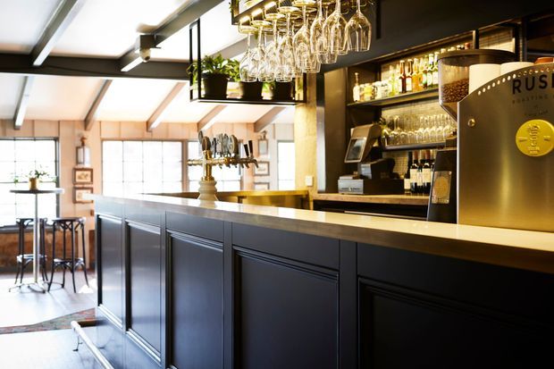 A warmly lit commercial bar interior with a long dark paneled counter, pale stone benchtop, beer taps, back-bar shelving, wine glasses, bottles, and a prominent beer dispensing unit. Industrial-style ceiling beams and glazing create a bright, inviting hospitality space, with small tables visible in the background.