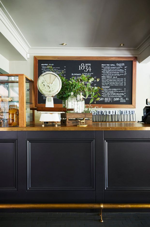 A refined café or bistro service counter viewed straight on. The foreground features dark charcoal-black paneled cabinetry with a warm timber countertop and brass foot rail. A framed chalkboard menu dominates the rear wall, accompanied by a glass pastry display, coffee equipment, jars, flowers, and small countertop items. The space has crisp white coffered-style ceiling trim, timber framing, and a polished, classic hospitality interior.