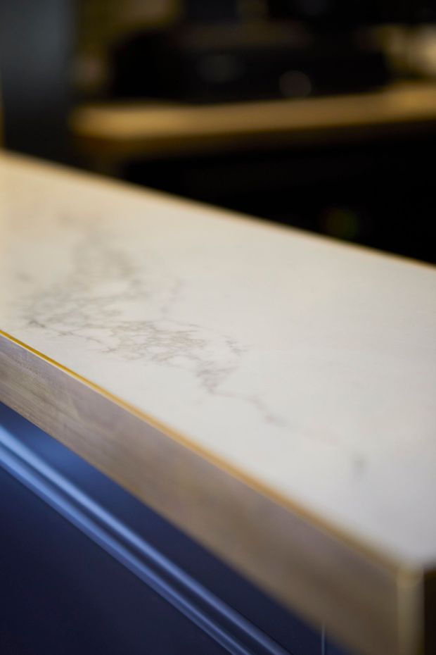 Close-up detail of a pale, veined stone countertop or benchtop with a softly rounded edge and a narrow warm-gold trim. The benchtop appears to be part of dark cabinetry or joinery, with a blurred dark interior background. The image is tightly framed and softly focused, emphasizing the surface material and edge detailing.