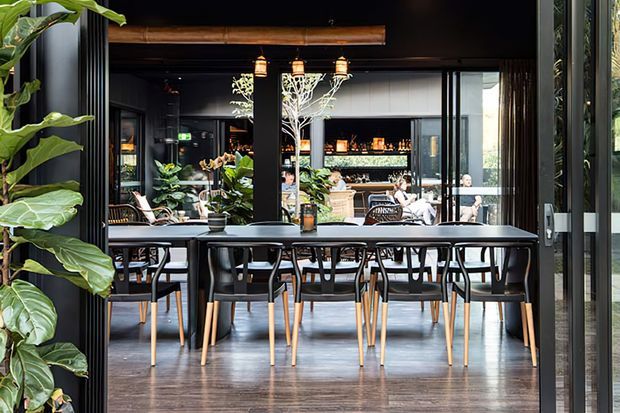 A dark, refined restaurant or hospitality dining space viewed through open glass doors. The foreground contains rows of timber-and-black dining tables and chairs, with pendant lights, potted plants, reflective glazing, and a bar or dining area visible beyond.