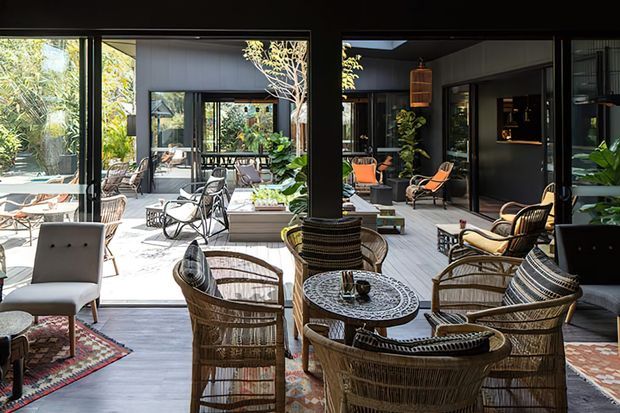 A bright, sophisticated indoor-outdoor living space arranged around a planted courtyard. The covered lounge features woven rattan armchairs and sofas, patterned cushions, a round patterned coffee table, dark structural columns, extensive black-framed glazing, and a tiled floor. Additional seating and lush greenery are visible across the courtyard, creating a relaxed resort-like atmosphere.