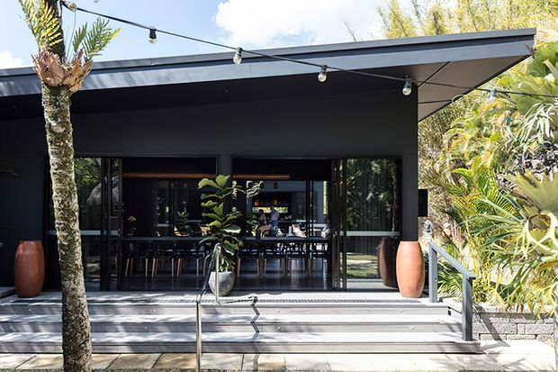 Exterior view of a contemporary dark-clad building opening onto a broad stepped terrace. Full-height glazed doors reveal a dining area inside, while tropical planting, large planters, steel handrails and festoon lighting frame the outdoor entry and entertaining space.