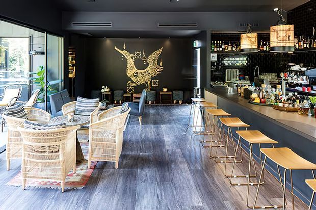 A stylish dark-toned hospitality interior featuring a long bar with timber stools, pendant lighting, extensive bottle shelving, lounge seating, and a dramatic gold bird mural on a black feature wall. Large windows bring in abundant daylight, contrasting with the charcoal finishes and warm timber accents.