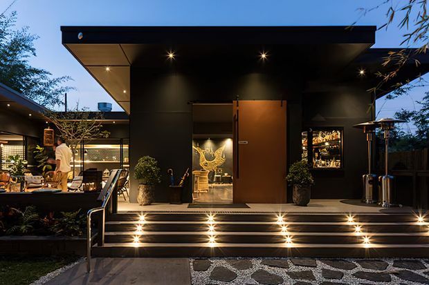 Twilight exterior view of a sophisticated modern pavilion-style residence or hospitality space, with dark cladding, broad flat roof overhangs, illuminated steps, a timber pivot door, and glazed openings revealing a warmly lit interior. Outdoor dining and lounge areas extend to the left, while a textured stone path and planted containers frame the entrance.