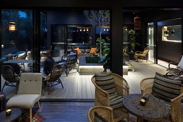 A dark, sophisticated indoor-outdoor entertaining area with extensive black-framed glazing opening onto a planted courtyard. The interior features woven rattan lounge chairs, a patterned round coffee table, a pale timber-look floor, and layered ambient lighting. Matching outdoor seating and potted greenery continue beyond the glazing, creating a seamless connection between the lounge and courtyard.