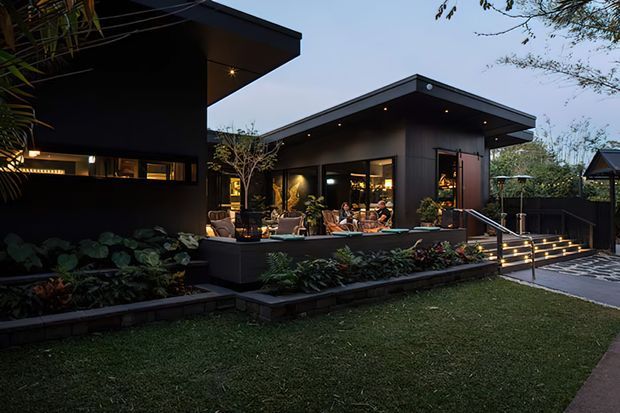 Twilight exterior view of a contemporary, low-profile pavilion-style residence with dark cladding, broad flat roof overhangs, warm recessed lighting, extensive glazing, and a landscaped terrace. Stepped garden beds, illuminated stairs, and a paved walkway lead toward an outdoor entertaining area.