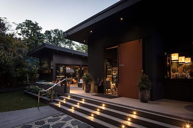 Twilight exterior view of a contemporary low-profile building with dark cladding, broad roof overhangs, illuminated steps, a timber-toned entry door, and landscaped surroundings. The open entry reveals a warmly lit interior and bar or reception area.
