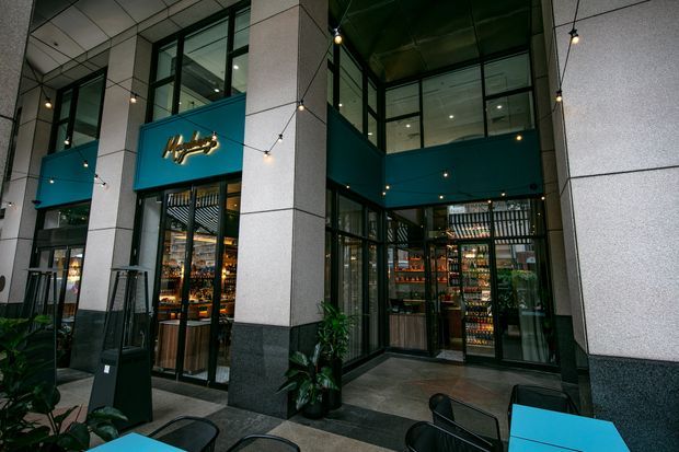 Street-level view of a contemporary commercial hospitality venue set within a tall glazed and stone-clad building arcade. The storefront has large black-framed windows and doors, teal feature panels, illuminated signage, visible bottle displays and bar shelving inside, plus outdoor café tables and chairs beneath suspended festoon lighting.