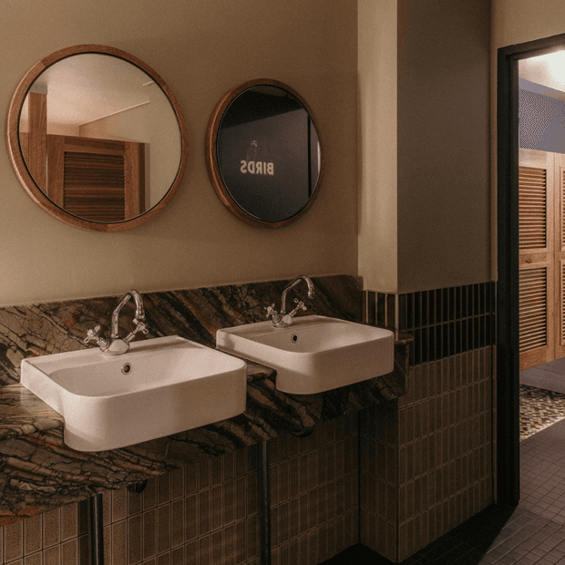 A warm, refined bathroom vanity with two wall-mounted white basins, chrome taps, a dramatic dark veined stone countertop and backsplash, and dark vertically gridded wall tiles. Round timber-framed mirrors hang above the basins, with a doorway visible to the side.