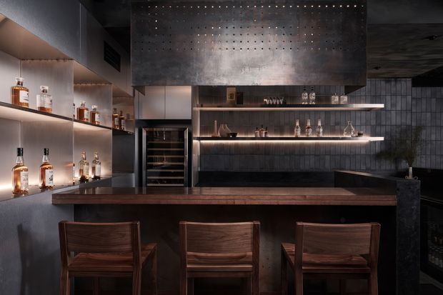 A moody, refined home bar with a dark timber counter and three matching timber bar stools. Backlit open shelving displays bottles and glassware, with a built-in wine refrigerator, dark textured wall tiles, and a large perforated metal range-hood-like feature above the bar area.