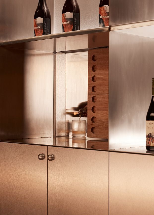 Close-up of a refined built-in bar or drinks cabinet with warm brushed metallic cabinetry, mirrored shelves and backing, integrated timber drawer fronts with circular finger pulls, and wine bottles displayed on the shelves. A decanter and tumbler are visible within the mirrored niche. The composition is warmly lit and highly reflective, with a luxe, tailored feel.