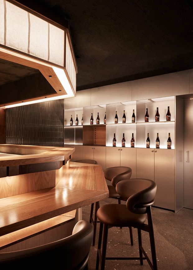 A warmly lit, sophisticated bar interior with a sculptural timber counter and tiered seating. Built-in illuminated shelving displays wine bottles against bronze-toned cabinetry, while dark textured wall and ceiling finishes create an intimate atmosphere. Curved brown bar stools line the counter, and a translucent illuminated overhead feature adds architectural interest.
