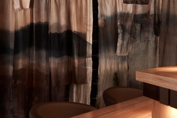 Close-up of a warm, moody interior detail featuring translucent, heavily textured dark brown and charcoal fabric or timber-like wall panels, with two rounded brown upholstered stools and a projecting warm timber counter or benchtop in the foreground.