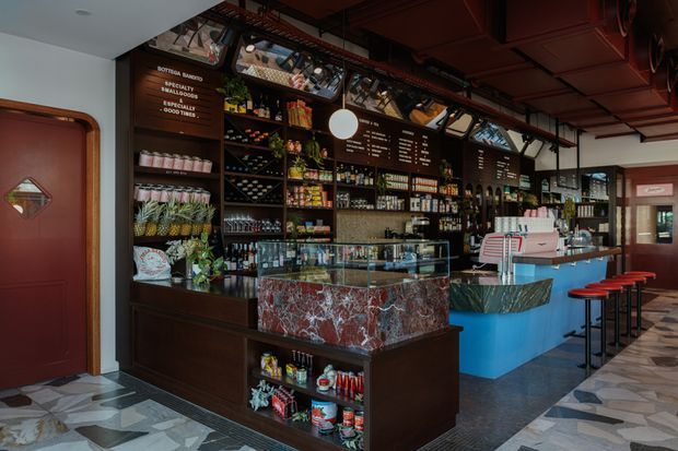 Interior view of a stylish café or bar with dark timber shelving, displayed beverages and pantry goods, a glass pastry or service counter, a colourful stone-clad island, blue cabinetry, red bar stools, menu boards, and patterned geometric flooring. The space has a rich, dramatic hospitality design with exposed dark ceiling services and distinctive decorative finishes.