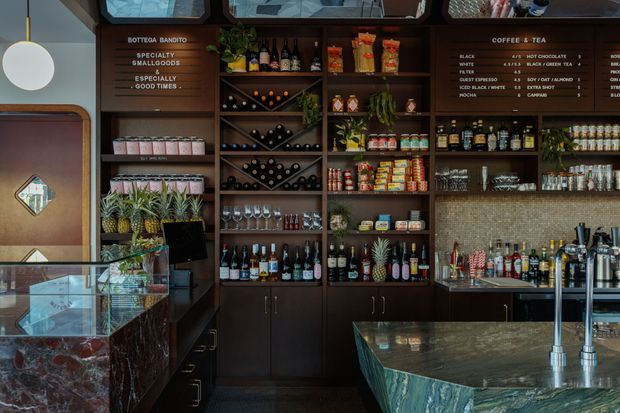 A richly detailed specialty coffee and food service counter interior, featuring dark timber shelving and cabinetry filled with coffee supplies, bottles, jars, glassware, and decorative plants. A large polished stone service counter with integrated taps occupies the foreground, while a coffee and tea menu is displayed above the shelving. The space has a refined, moody hospitality aesthetic with warm accent lighting and patterned tile backing.