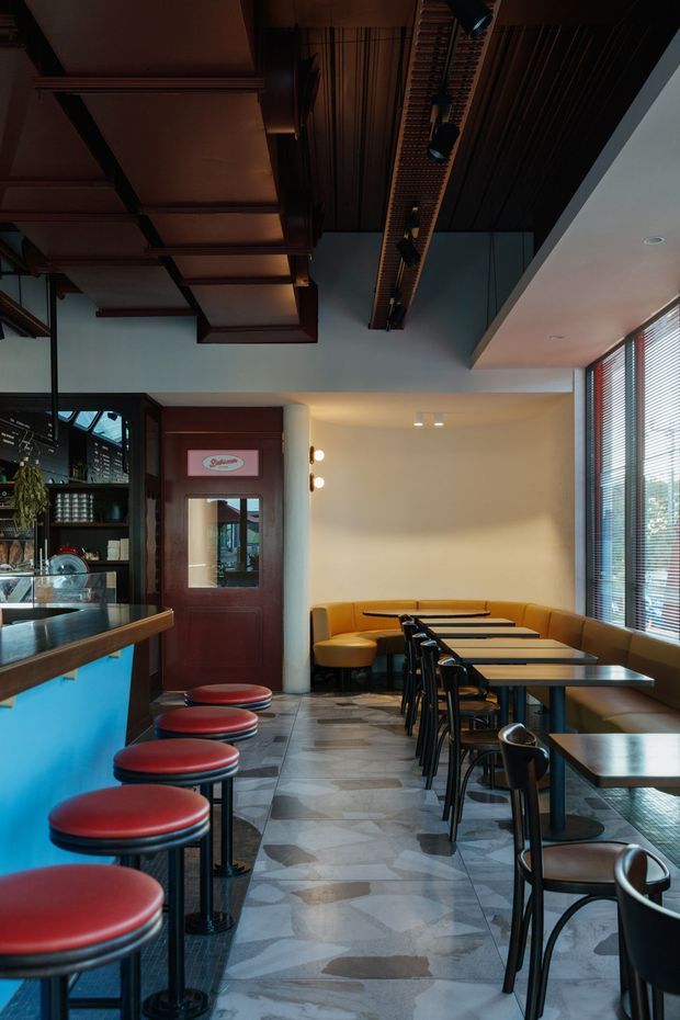 Interior view of a compact retro-styled café or diner, with a long counter and red upholstered stools on the left, rows of dark tables and chairs, and a curved mustard banquette beneath large windows with horizontal blinds. The space features dark timber ceiling detailing, a patterned stone or tile floor, warm cream walls, and a bright blue counter front.
