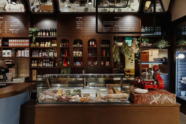 A warmly lit deli and specialty food shop interior with dark timber wall cabinetry, arched display niches, open shelving stocked with packaged goods and bottles, hanging plants, overhead menu boards, a large refrigerated glass food counter, and a red commercial meat slicer on a marble-patterned countertop. The space has a refined, richly layered hospitality-retail fit-out.