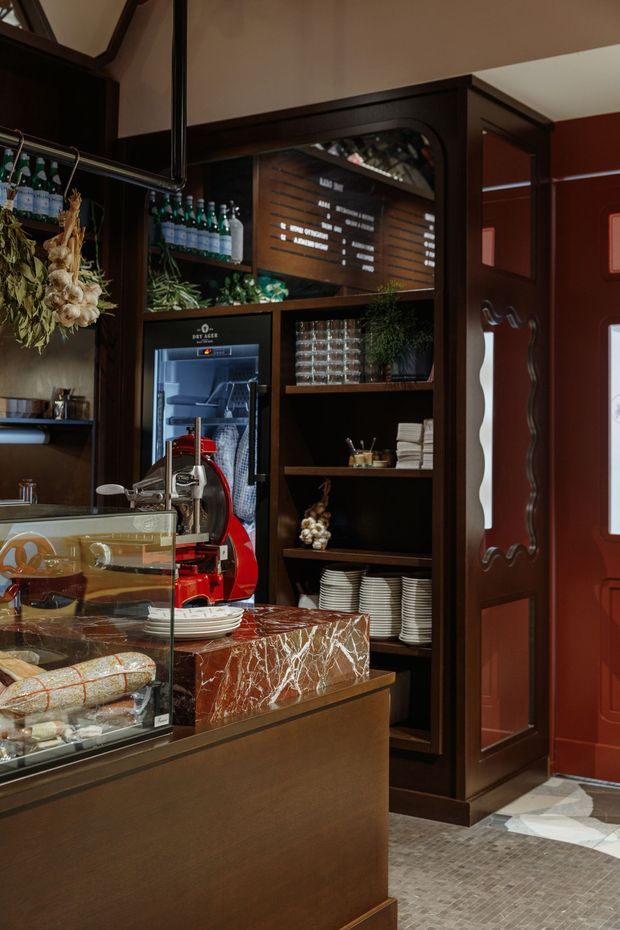 A richly detailed café or bar interior with dark timber shelving and cabinetry, stocked with bottled drinks, glassware, plates, and small decorative displays. A marble service counter with a glass pastry case and coffee equipment occupies the foreground. A deep red paneled door, patterned stone or tile flooring, hanging dried botanicals, and a backlit menu board contribute to an upscale, moody atmosphere.