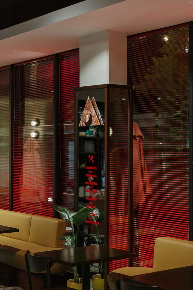 A moody contemporary commercial interior with a café or lounge seating area. Floor-to-ceiling glazing is fitted with vivid red horizontal blinds, creating strong linear contrast and reflections. Tan upholstered sofas and armchairs surround dark timber tables, while a black-framed illuminated display or partition features artwork and red lettering. Potted plants soften the dark palette, and a white ceiling with recessed lighting completes the space.