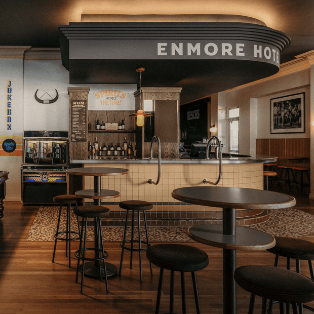A warmly lit retro-inspired hotel bar with a prominent curved black canopy sign reading “ENMORE HOTEL.” The space features a tiled rounded bar counter with integrated taps, dark round stools and pedestal tables, timber flooring, patterned rugs, vintage-style spirits shelving, a jukebox, wall panelling, framed artwork, and decorative pendant lighting.