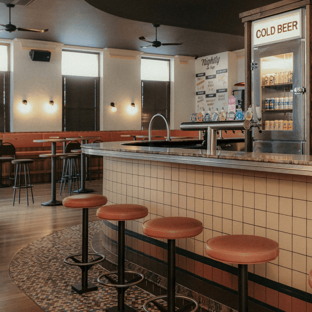 A retro-inspired commercial bar interior with a tiled counter, row of red upholstered stools, brass-toned trim, beer taps, and a glass-front cold beer cabinet. The space includes café-style tables and stools, booth seating, ceiling fans, wall sconces, and vintage graphic signage, creating a warm mid-century diner or pub atmosphere.