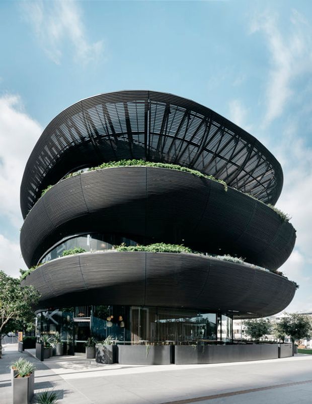 Exterior view of a striking multi-storey contemporary building with rounded, stacked floor plates and extensive dark horizontal metal screening. Curved rooftop and terrace structures incorporate lush linear planting, while the ground level has extensive glazing, outdoor planters, and a paved urban forecourt. The building is photographed from a low angle against a bright sky.