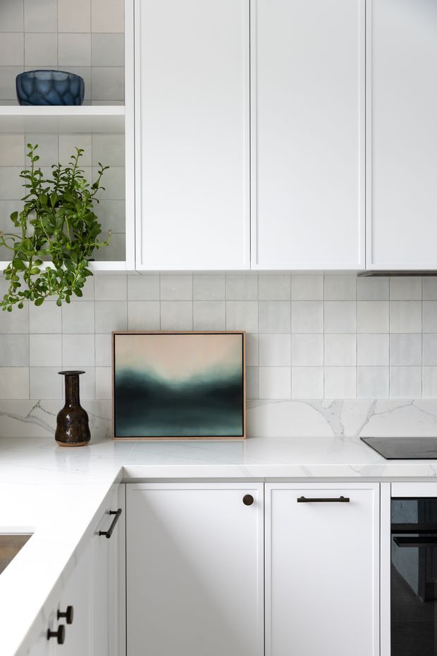 A bright, refined kitchen interior featuring white shaker-style cabinetry, a pale tiled backsplash, and a white marble-look benchtop. A dark cooktop and oven are partially visible at right, while open shelving displays a blue vessel and greenery. A framed abstract artwork and dark decorative vase add contrast to the predominantly white scheme.
