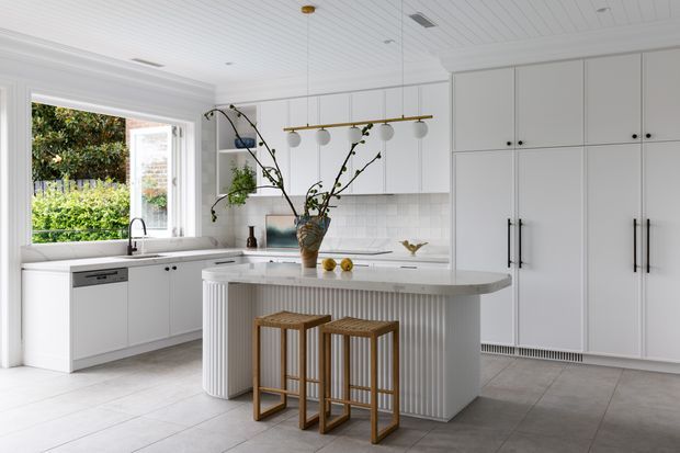 Bright, refined white kitchen with a large curved island featuring vertically fluted detailing, integrated appliances, tall shaker-style cabinetry, a tiled backsplash, and a timber-accented pendant light. Two timber stools sit at the island, while large windows bring in garden views and natural light.
