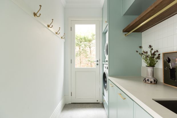 A bright, compact laundry room with pale sage-green cabinetry, a white benchtop, integrated front-loading washing machine and dryer, and a deep black sink. Open timber shelves with brass supports run above the counter, while a row of brass wall hooks lines the white wall. A glazed exterior door brings in natural light, and simple dried botanicals add a small decorative accent.
